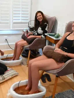 Three women sitting in pedicure chairs in a bright spa room, smiling during a foot soak.