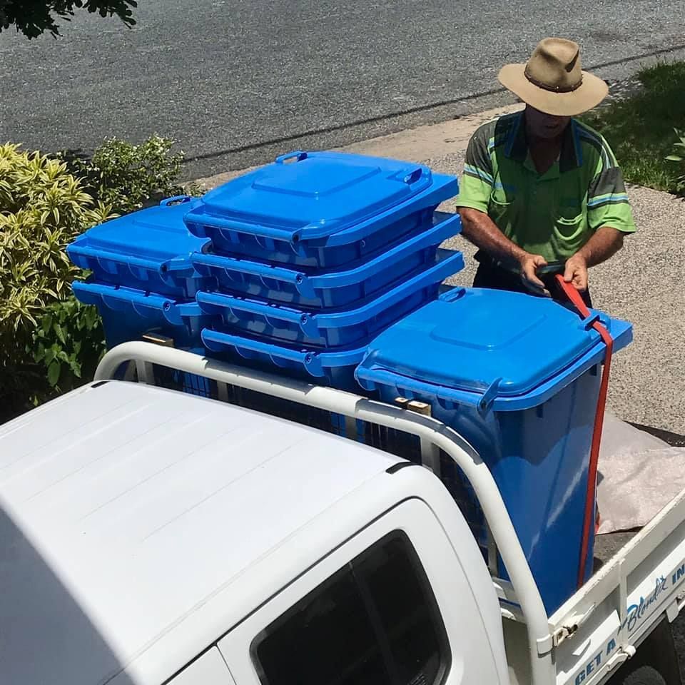 A Man In A Hat Is Loading Blue Garbage Cans Into A Truck — Blondies Bins in Rural View, QLD