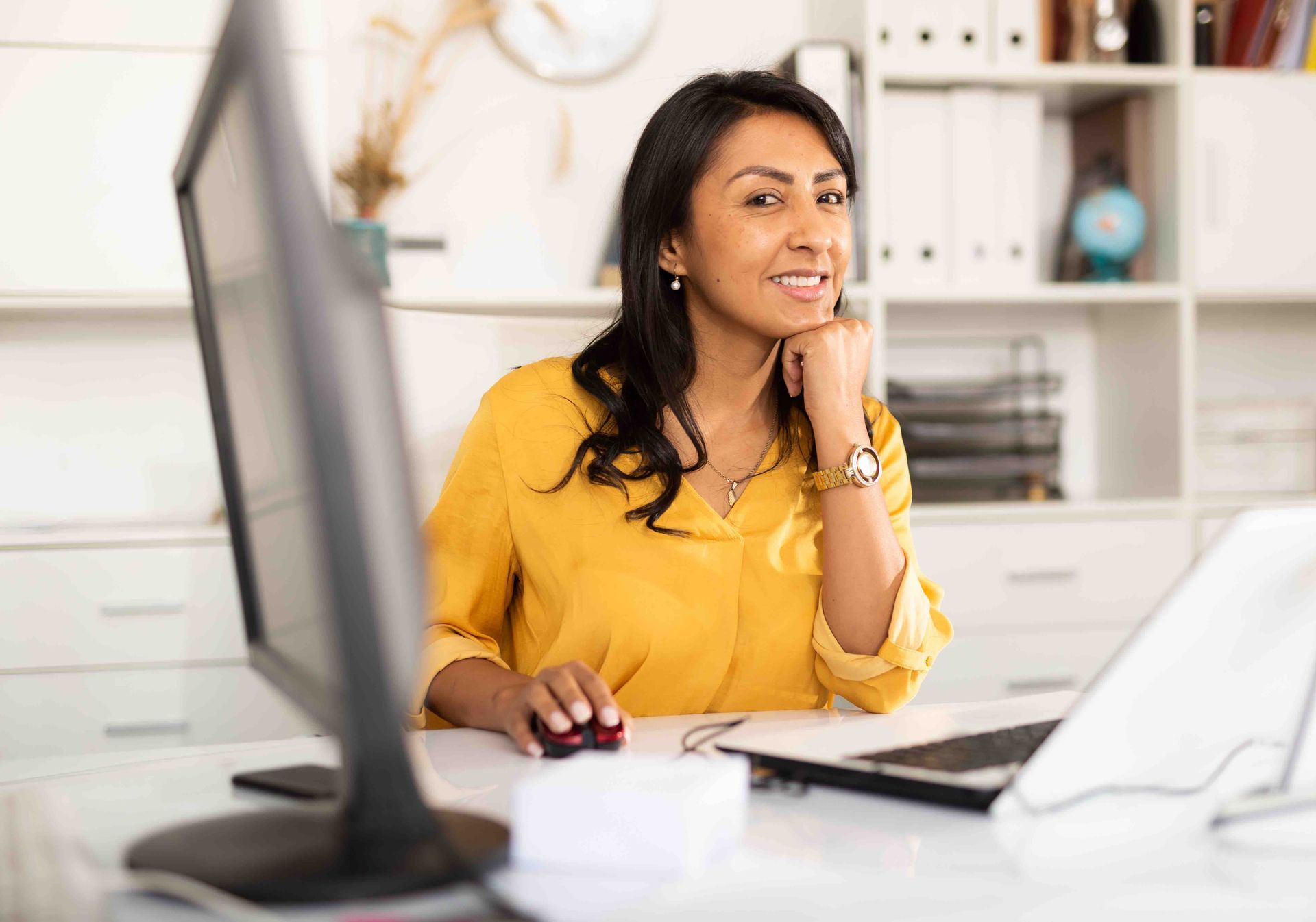 Woman smiling at her desk with a computer and laptop, wearing a yellow shirt.