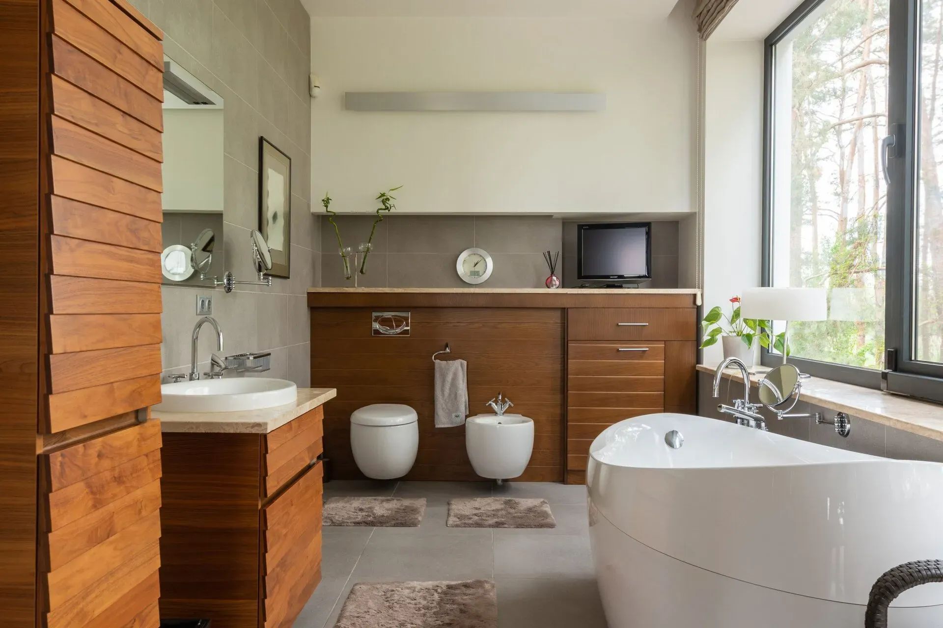 Modern bathroom with wood cabinetry, twin toilets, a freestanding tub, and a large window