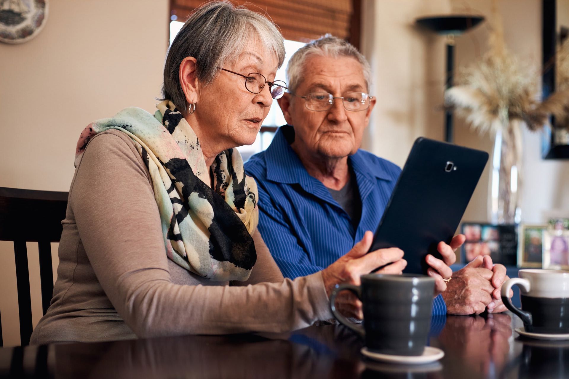 An elderly couple is sitting at a table looking at a tablet.