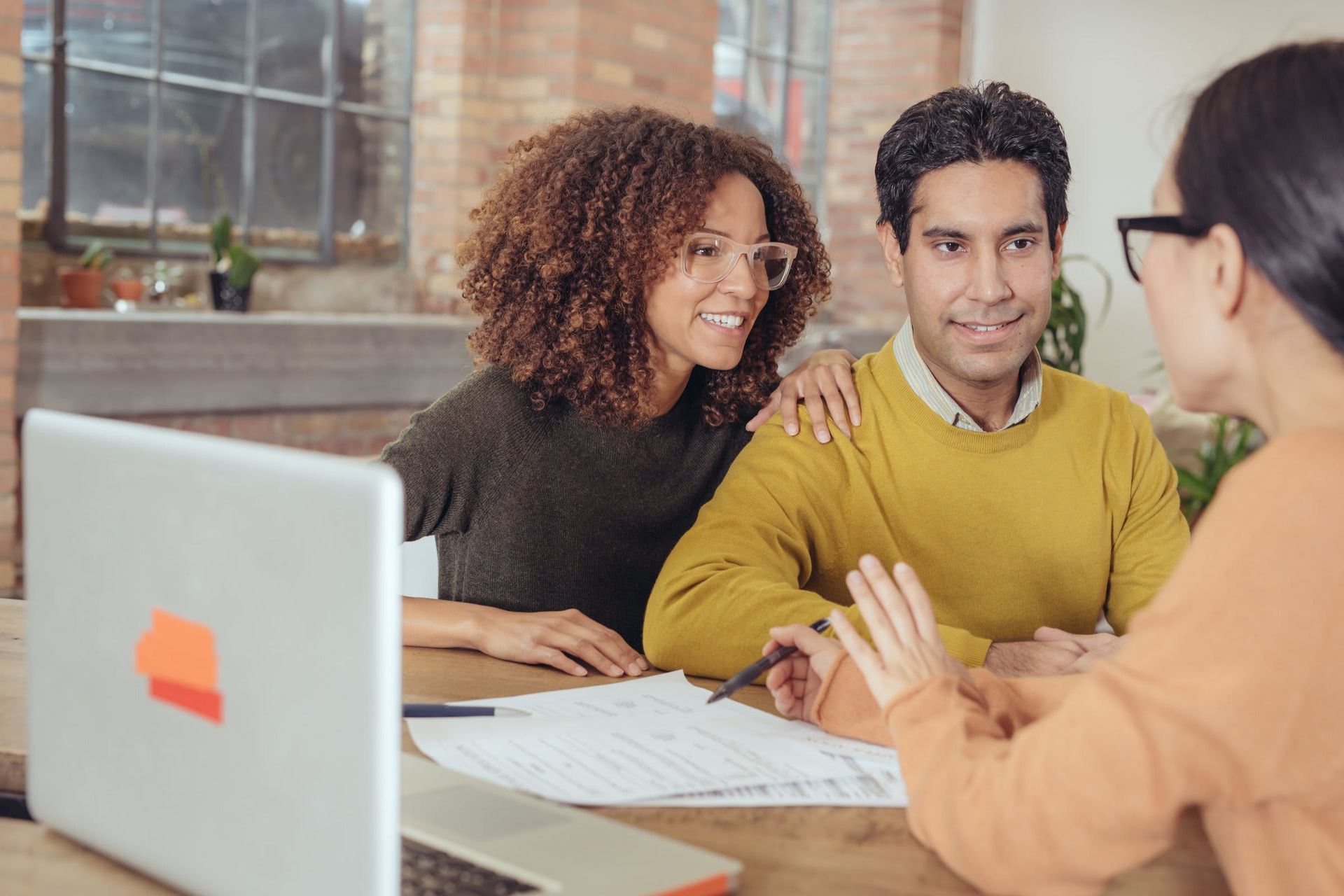 A man and woman are sitting at a table talking to a woman in front of a laptop.
