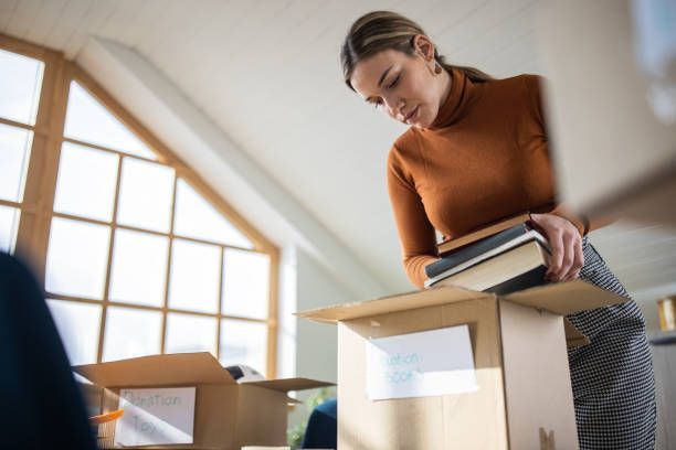 A woman is decluttering, packing toys and books into a cardboard box before moving services.