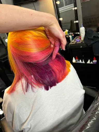 A hand lifts a section of vibrant, multi-colored hair showing layers of yellow, orange, bright pink, and deep purple.