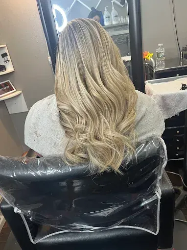 A person sits in a salon chair, facing away from the camera, showcasing long, wavy, highlighted blonde hair.