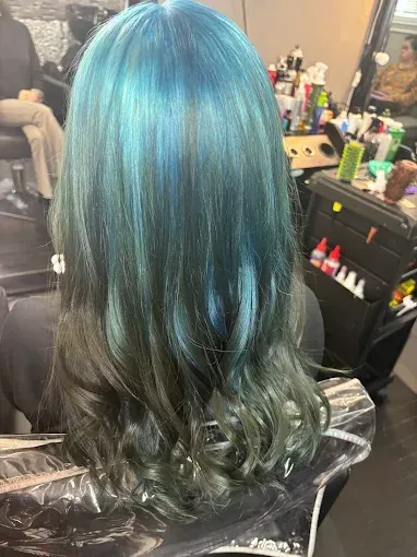A person sitting in a salon chair with long wavy hair dyed bright turquoise at the roots, fading into dark forest green.