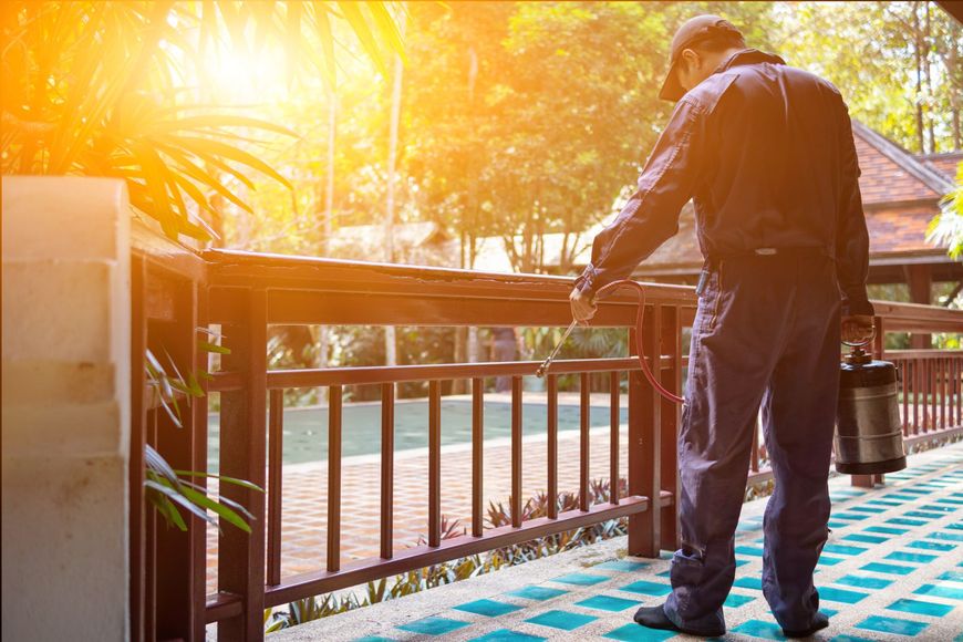 Pest control worker spraying a wooden railing on a patio, holding a tank. Bright sunlight.