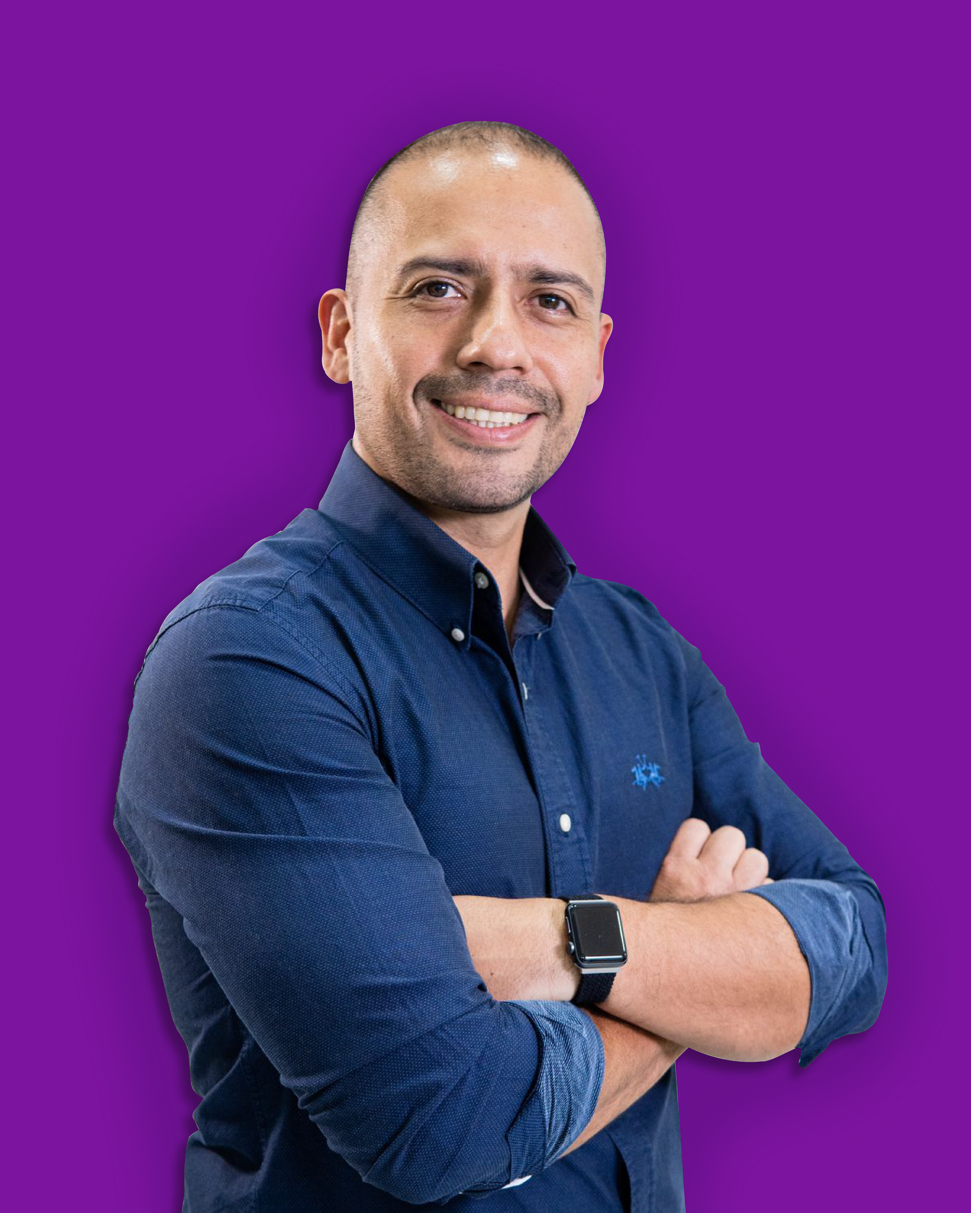 Hombre con los brazos cruzados sonriendo, vistiendo una camisa azul abotonada, fondo morado.