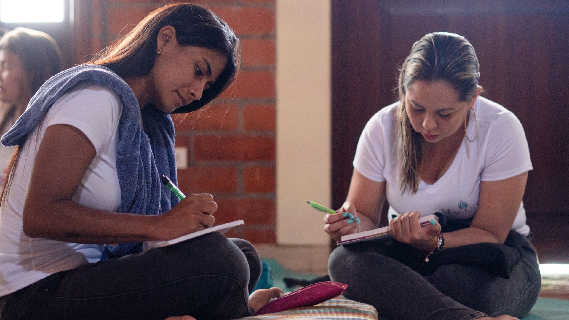 Dos mujeres sentadas con las piernas cruzadas, escribiendo en cuadernos. Una lleva una camisa blanca, la otra tiene una toalla azul.