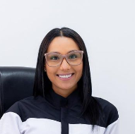 Mujer con gafas sonríe, sentada en una silla. Lleva una blusa blanca y negra, en el interior.