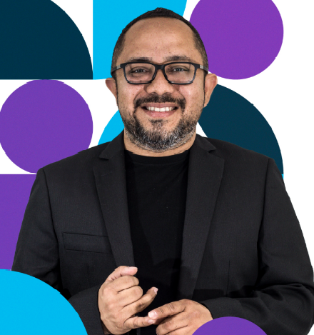 Hombre con gafas sonriendo, con chaqueta negra, sobre un fondo geométrico morado y azul.