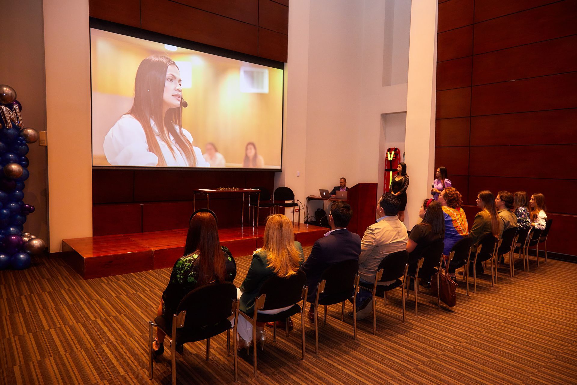 El público observa una gran pantalla donde aparece una mujer hablando. Las personas están sentadas en sillas en una sala moderna.