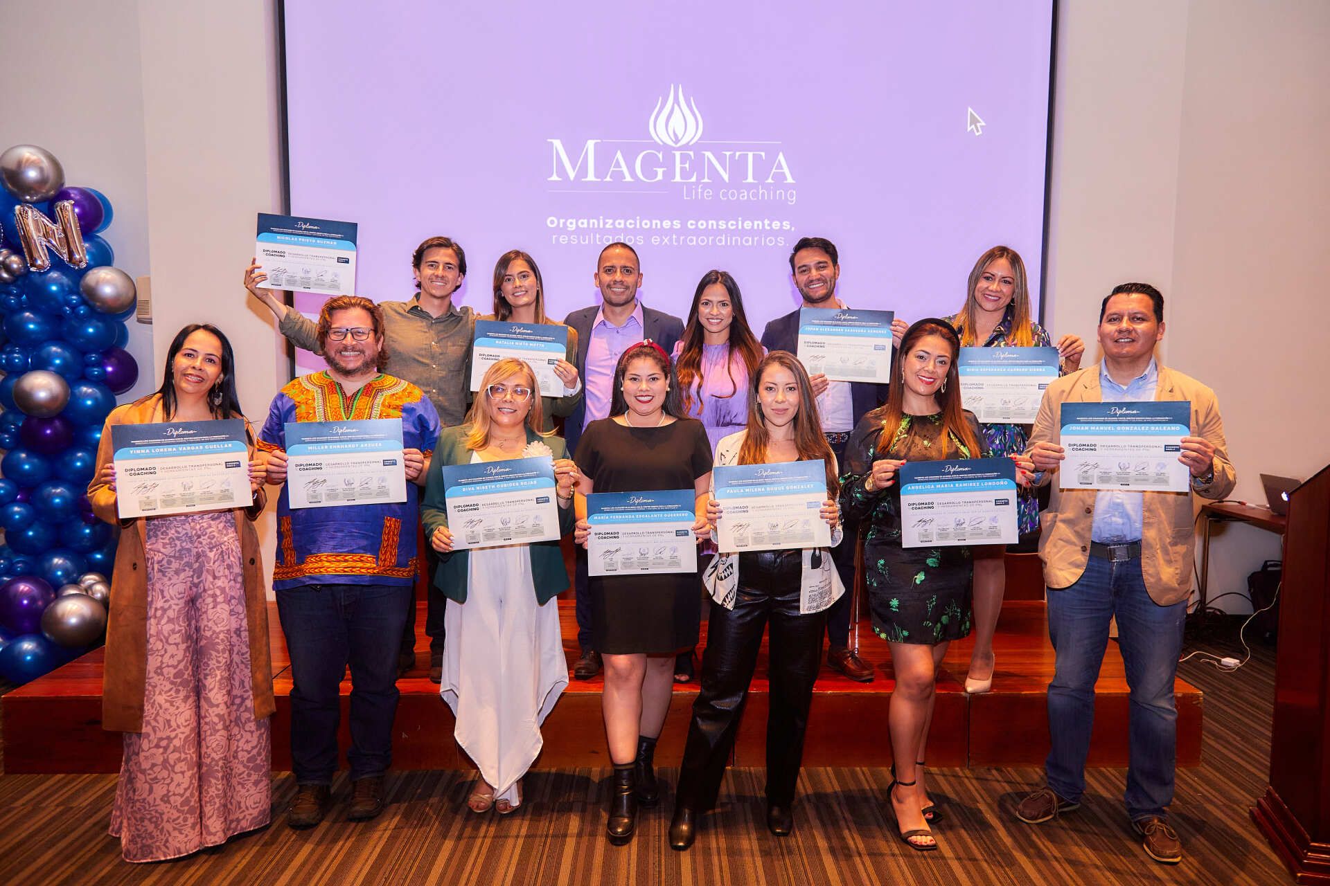 Grupo de personas sosteniendo certificados, sonriendo, en un interior con un fondo morado que dice “Magenta”.
