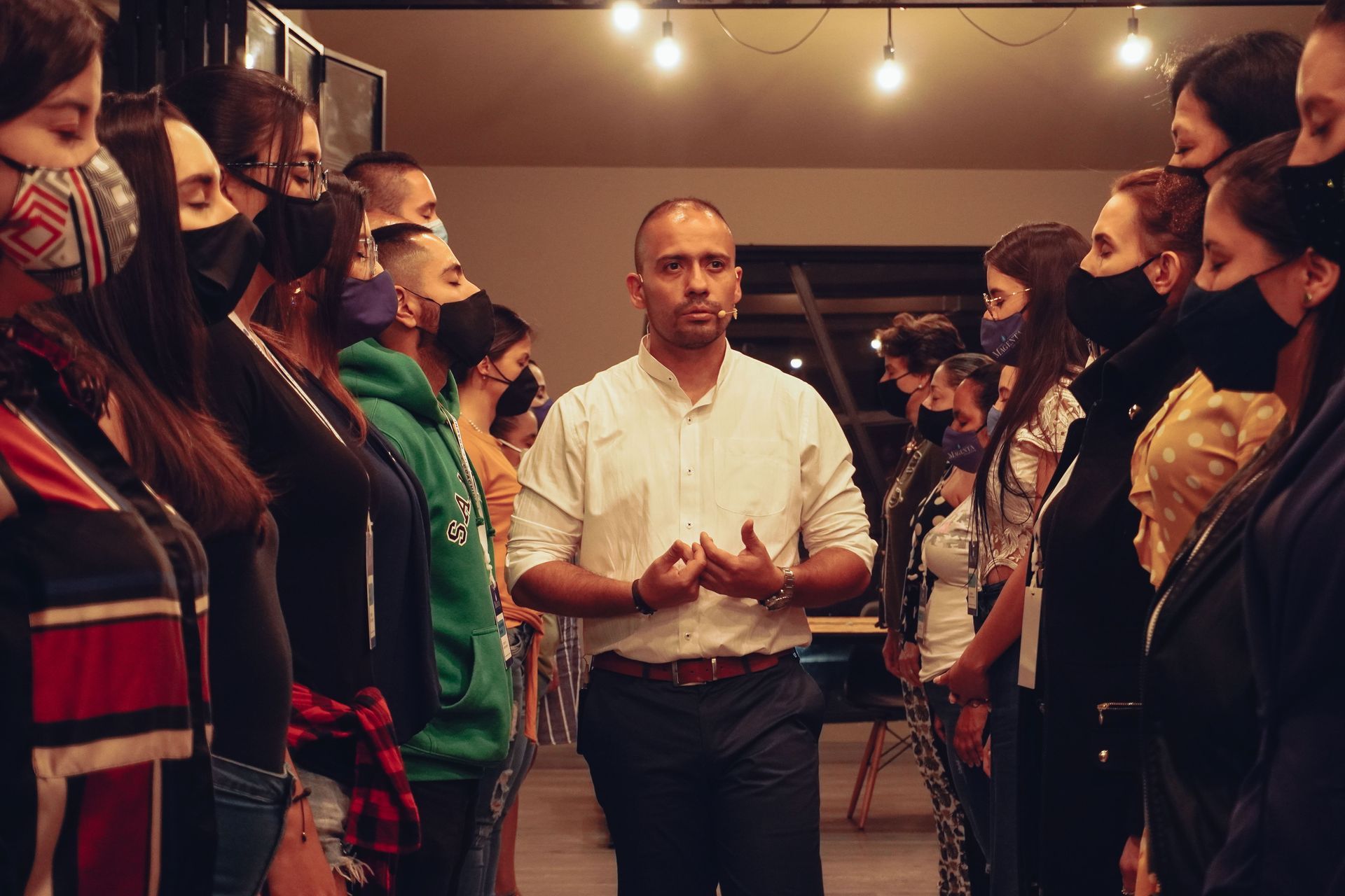 Un hombre con las manos en alto habla a un grupo de personas que llevan mascarillas, en un espacio interior iluminado con guirnaldas de luces.