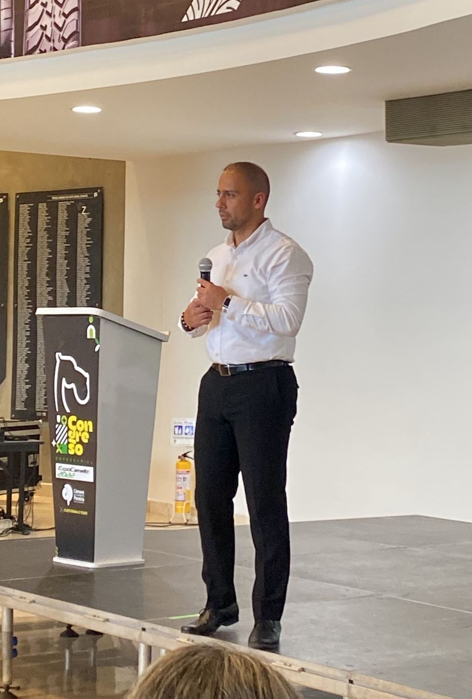 Hombre hablando desde un podio, sosteniendo un micrófono. Lleva camisa blanca y pantalones negros, en un salón.
