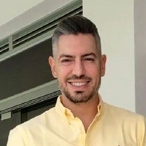 Hombre con canas sonriendo, vestido con una camisa amarilla de cuello. De pie frente a una ventana.