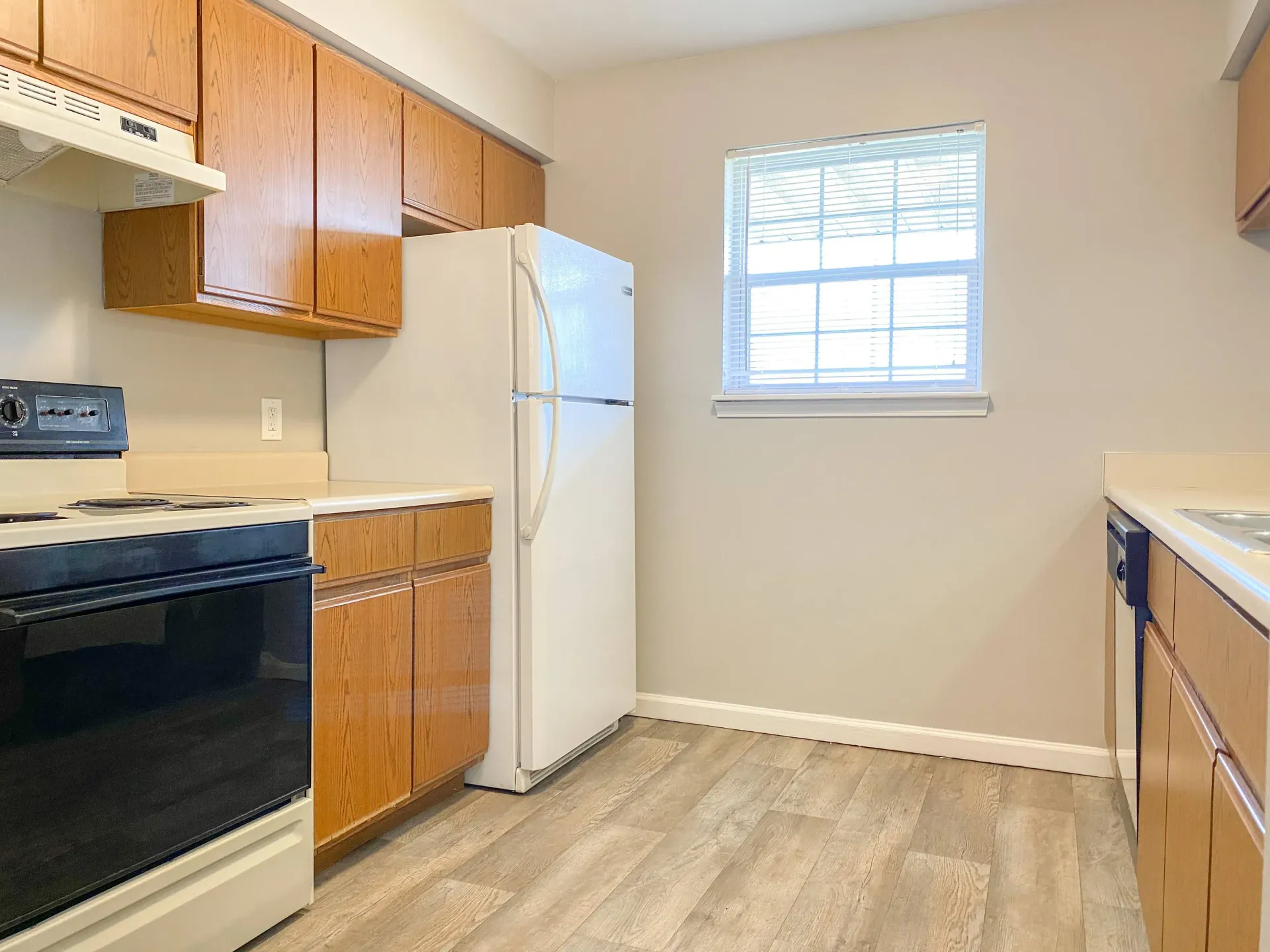 Apartment kitchen with wooden cabinets, white fridge and stove, and a window with blinds.