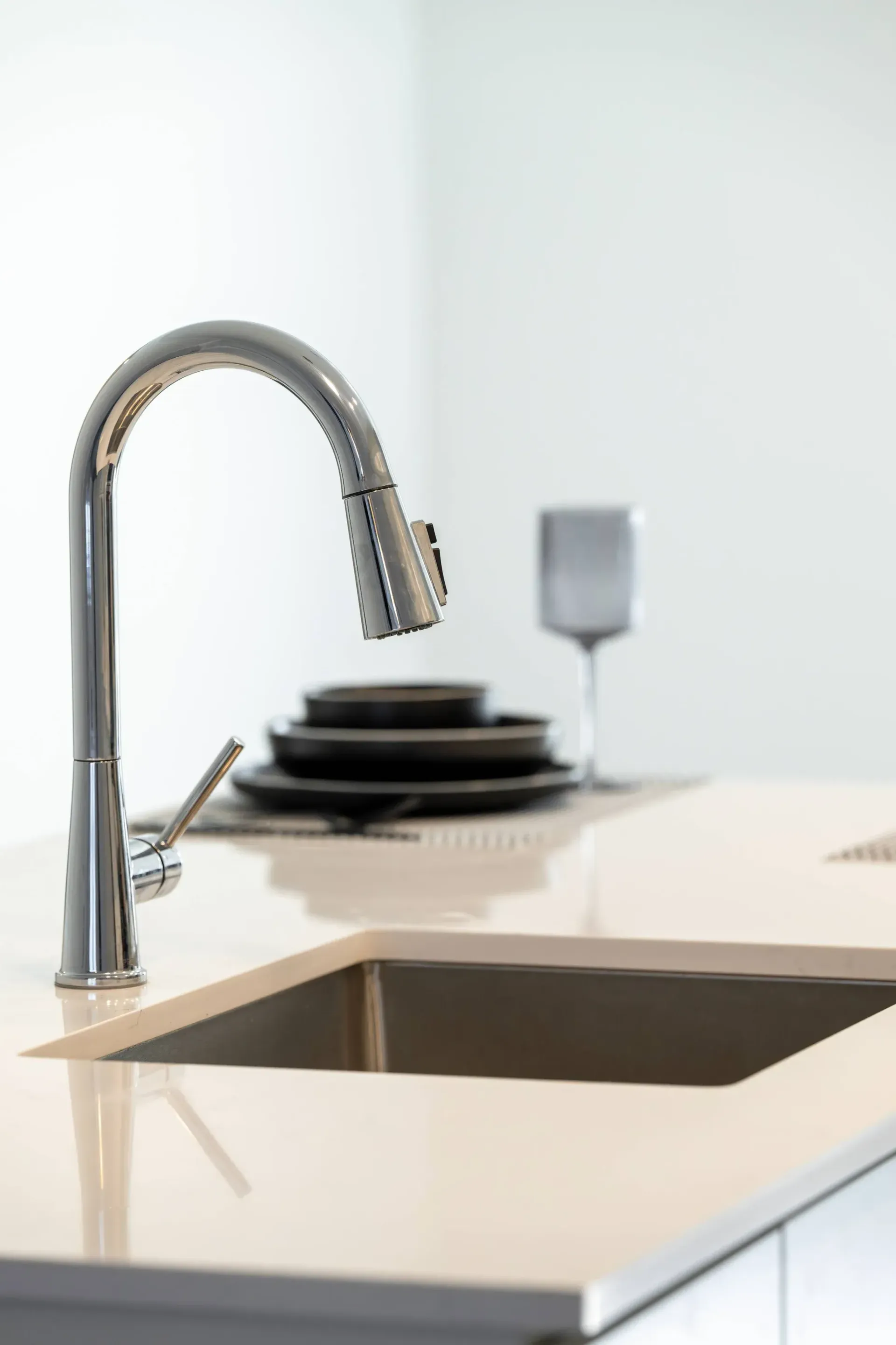 Close-up of a chrome kitchen faucet over a sink with stacked plates and a wine glass in the background.