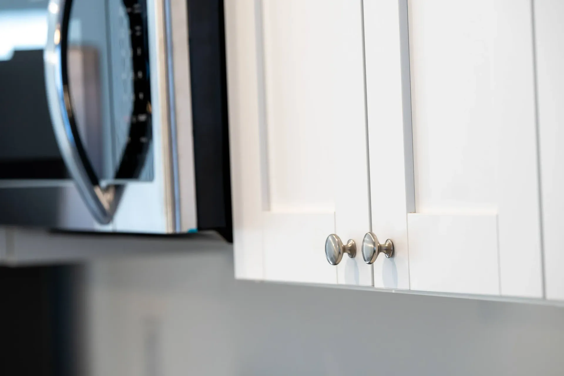 Close-up of white kitchen cabinetry with silver knobs and a stainless microwave on the left.