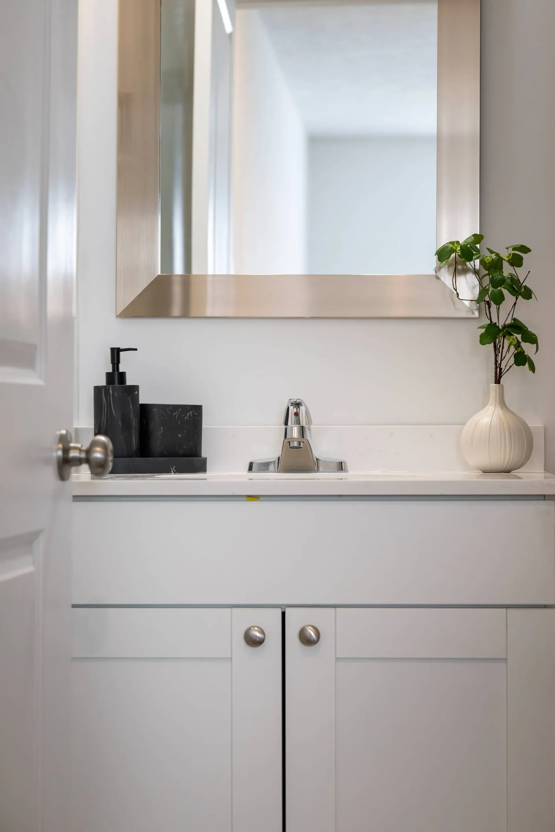 White bathroom vanity with marble countertop, chrome faucet, and a plant by a large framed mirror.