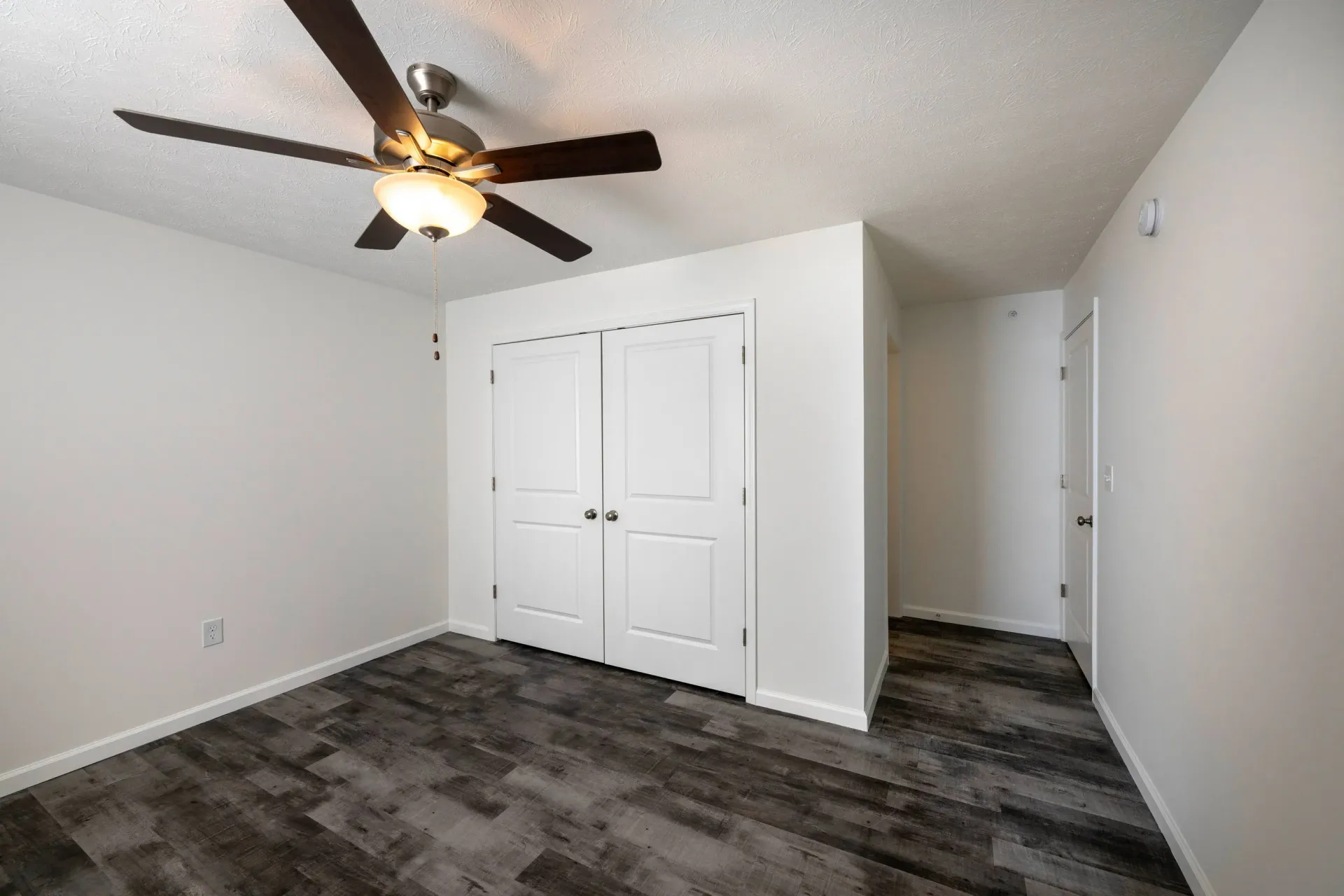 Bedroom with white walls, dark wood-like flooring, ceiling fan, and double closet doors.