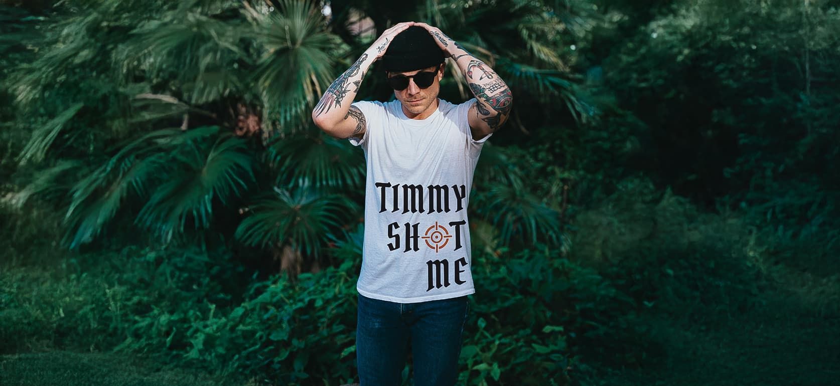 A man amongst foliage wearing a white t-shirt with Timmy Shot Me Logo Design on it
