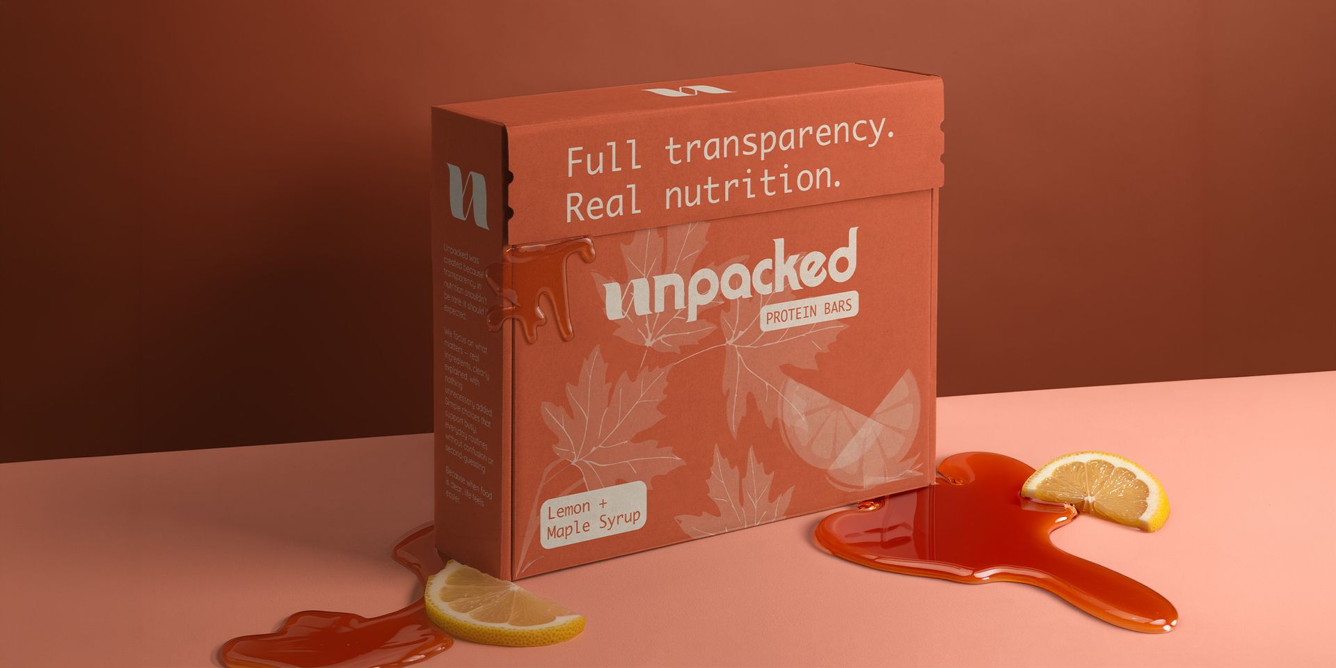 A box of Unpacked brand protein bars on a green surface with lemon slices and maple syrup oozing from the box.