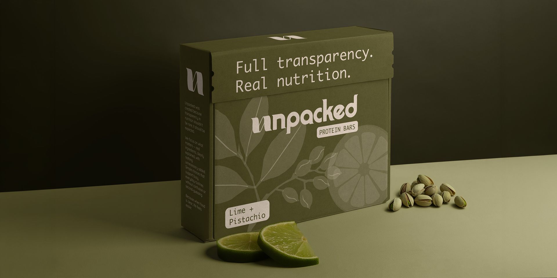 A box of Unpacked brand protein bars on a green surface with lime slices and a small pile of pistachio around the box.