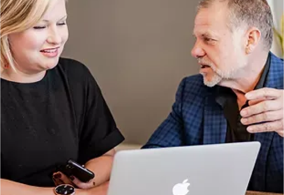 A man and a woman are sitting at a table looking at a laptop computer.