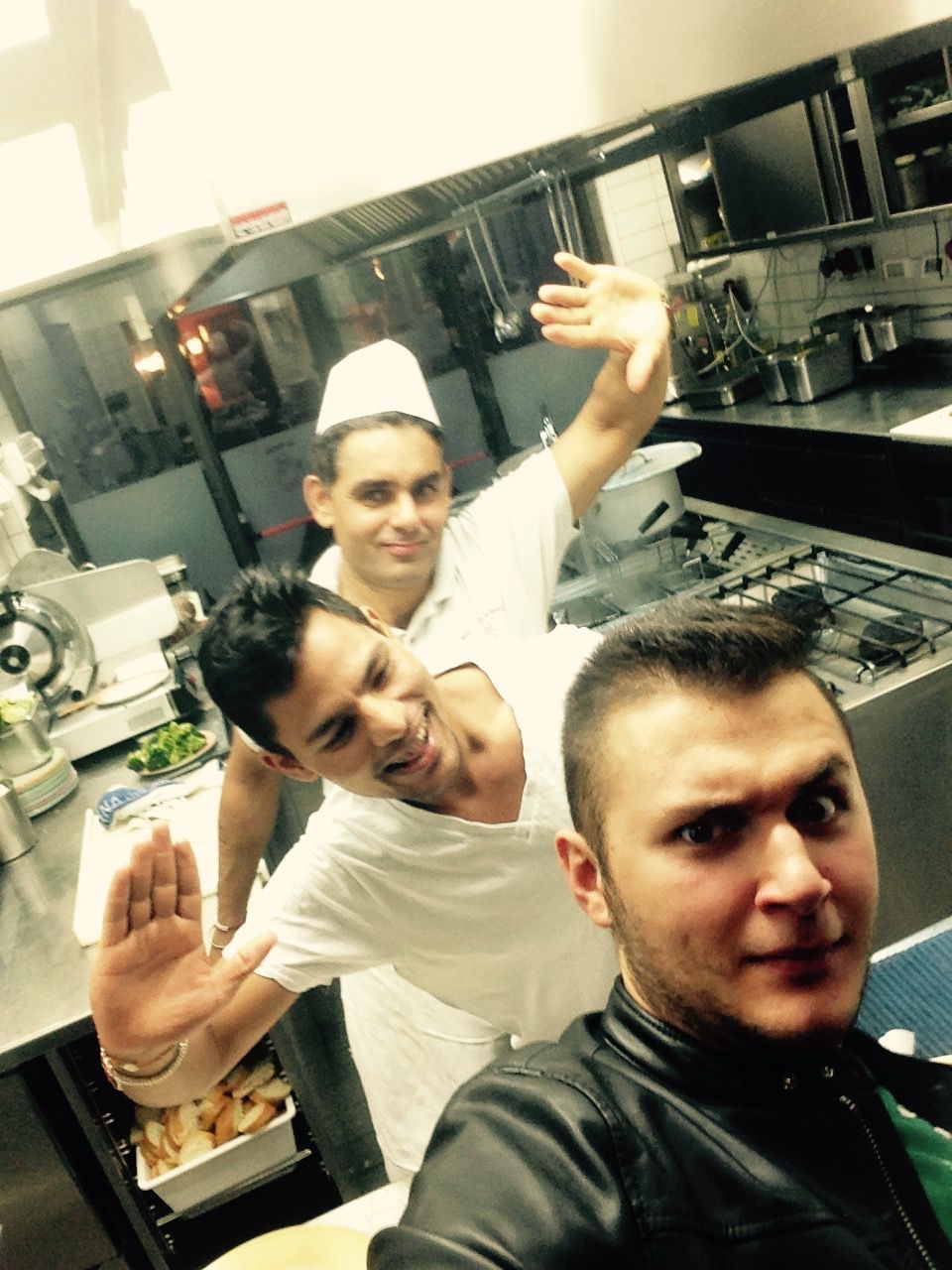 Tre dipendenti di un ristorante si scattano un selfie in cucina. Uno indossa un cappello da chef, sorride e saluta.
