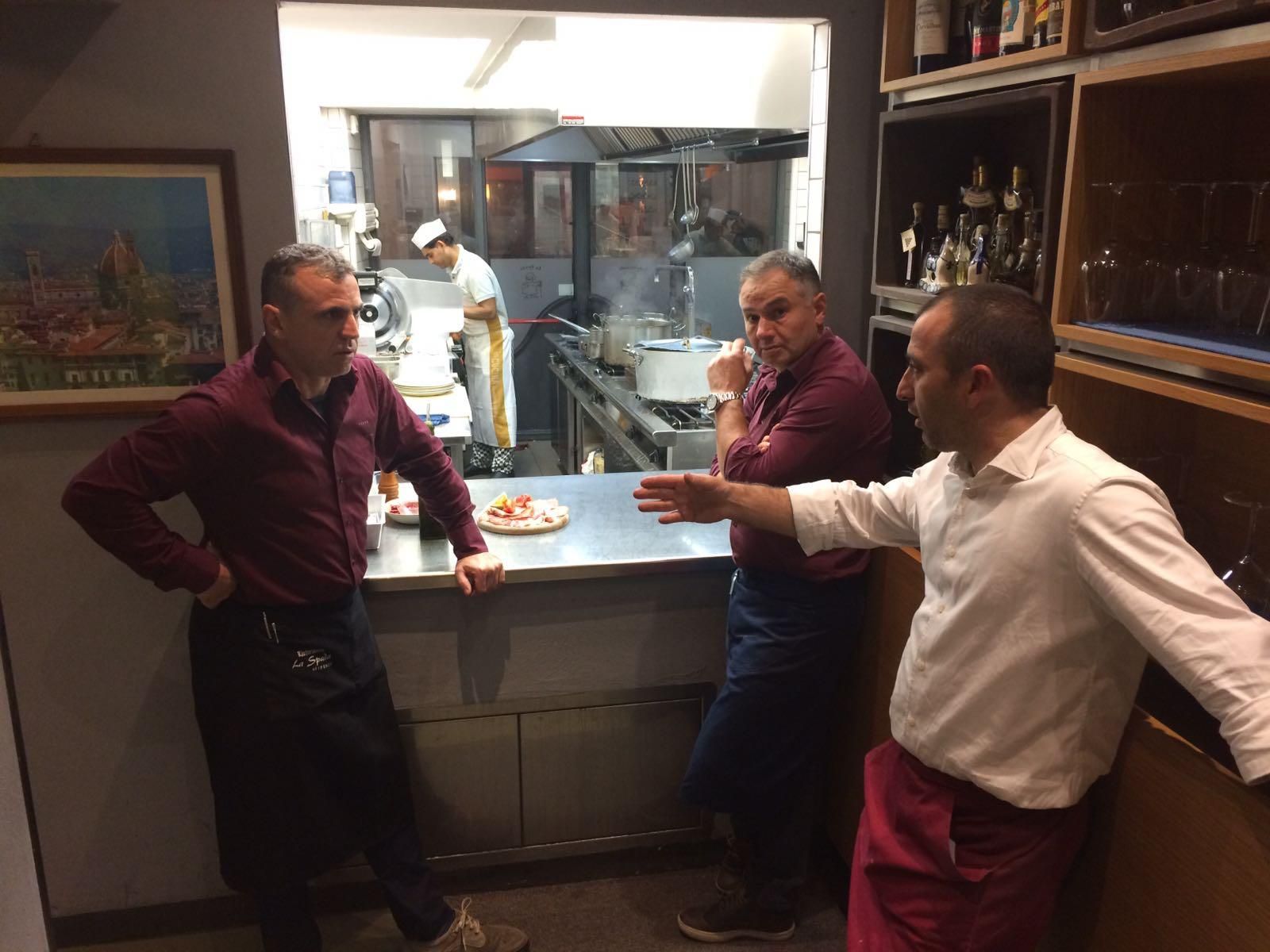 Tre uomini in un ristorante discutono vicino alla cucina. Uno di loro fa un gesto, lo chef è sullo sfondo.