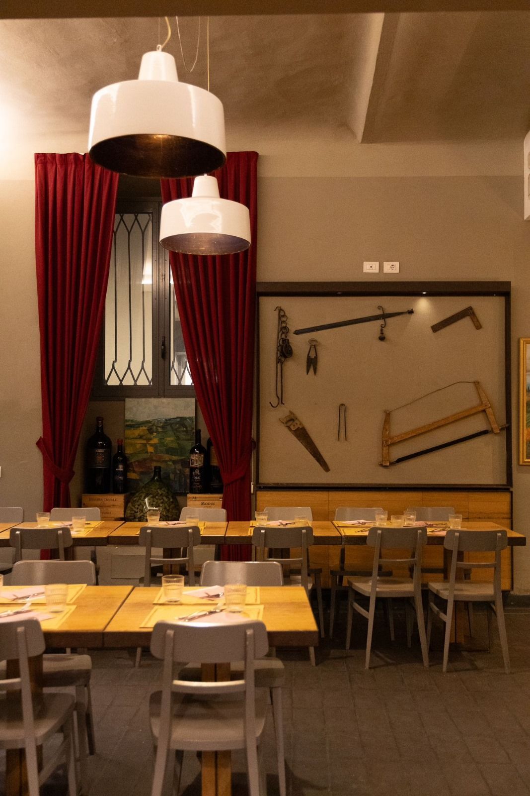 Interno del ristorante con tavoli, sedie, tende rosse e lampade bianche appese. Attrezzi esposti alla parete.