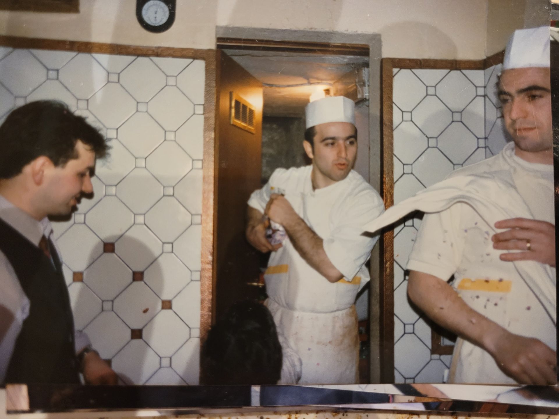 Tre uomini in cucina, due in divisa da chef. Uno si esamina le mani mentre l'altro gesticola. Paraschizzi piastrellato.