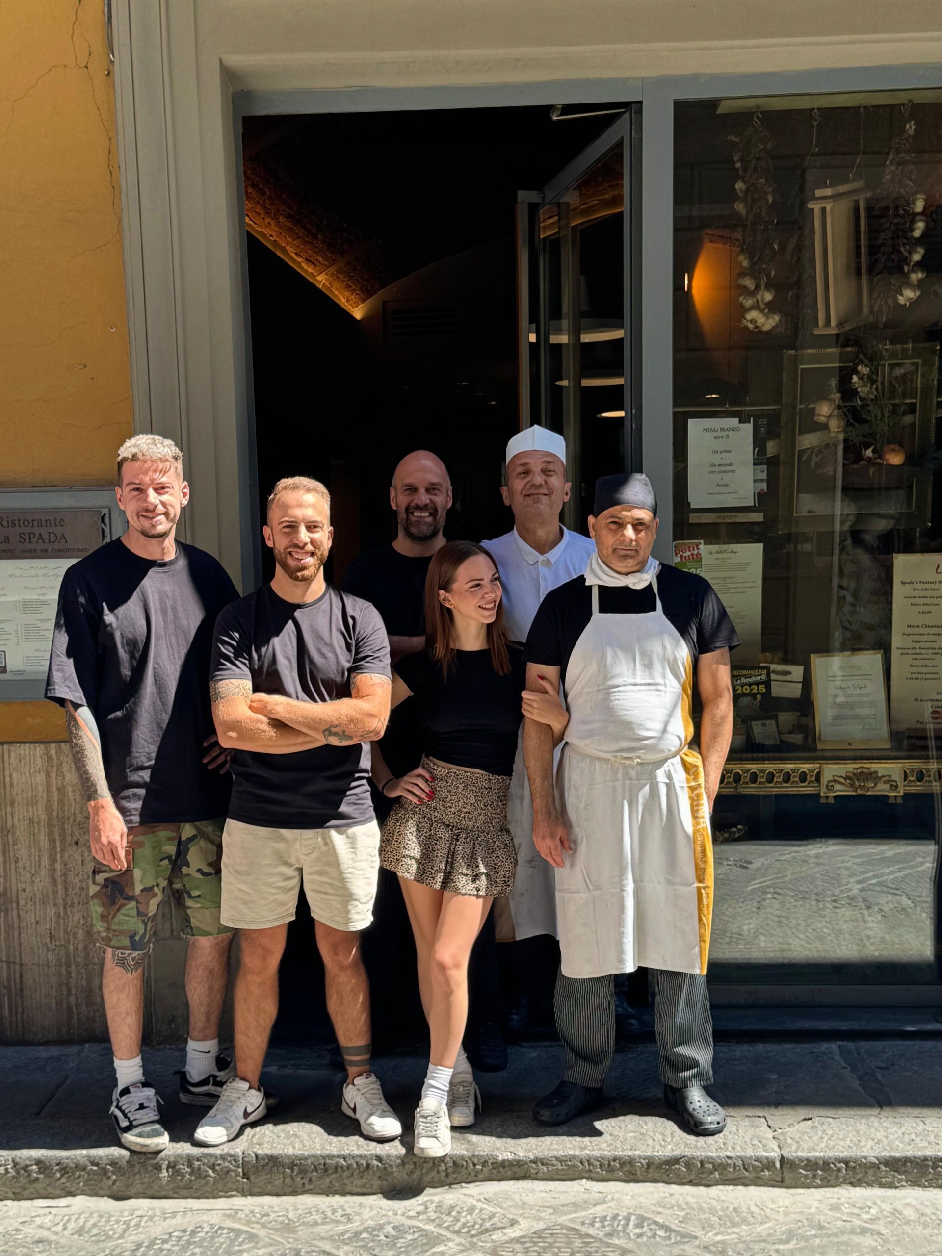 Tre donne sorridenti, a braccetto, che guardano la telecamera. Una indossa un'uniforme nera con un logo.