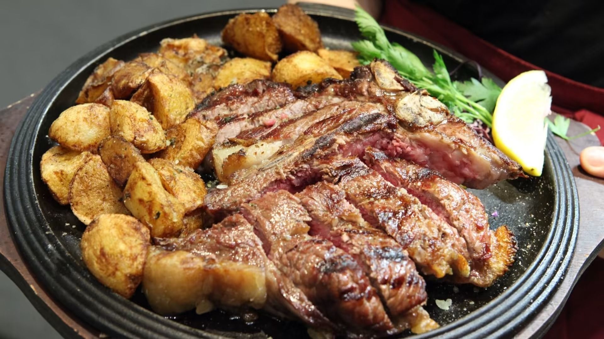 Bistecca alla griglia tagliata a fette con patate arrosto su un piatto sfrigolante, guarnita con prezzemolo e limone.