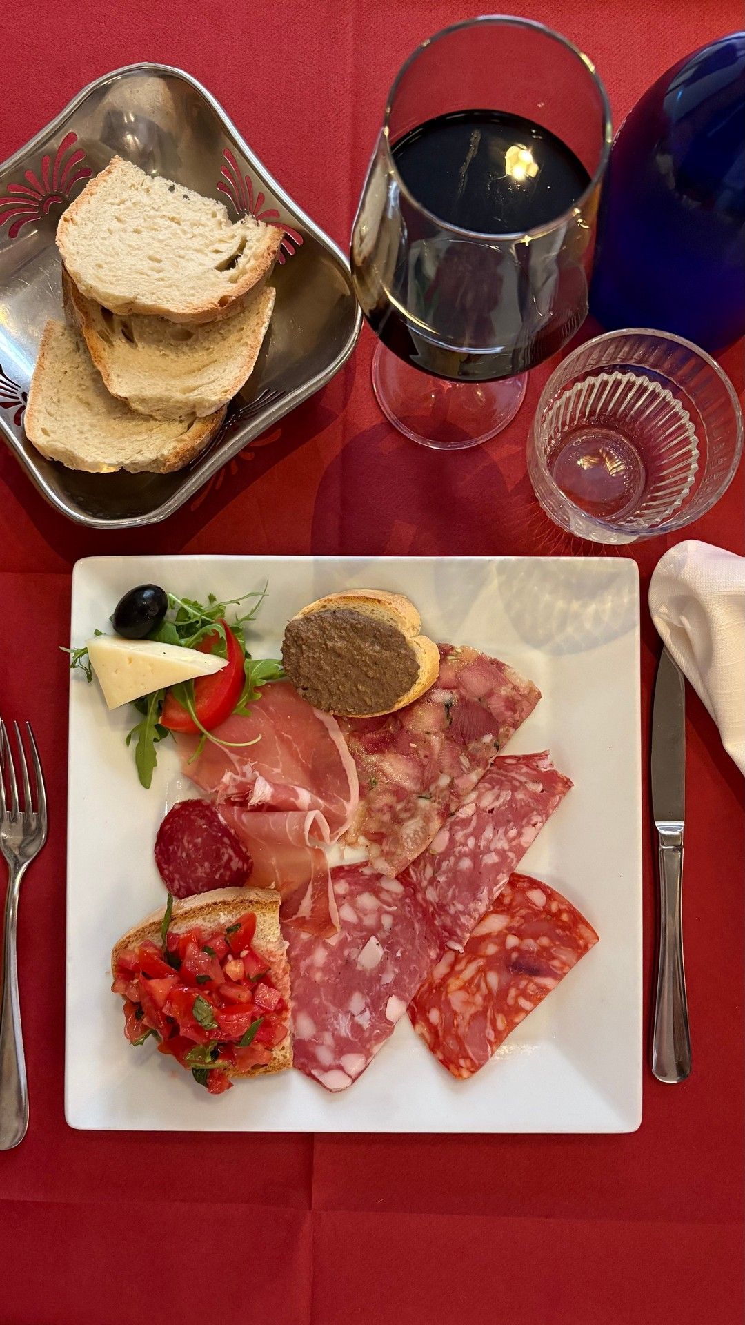 Piatto di salumi, pane e vino su una tovaglia rossa.