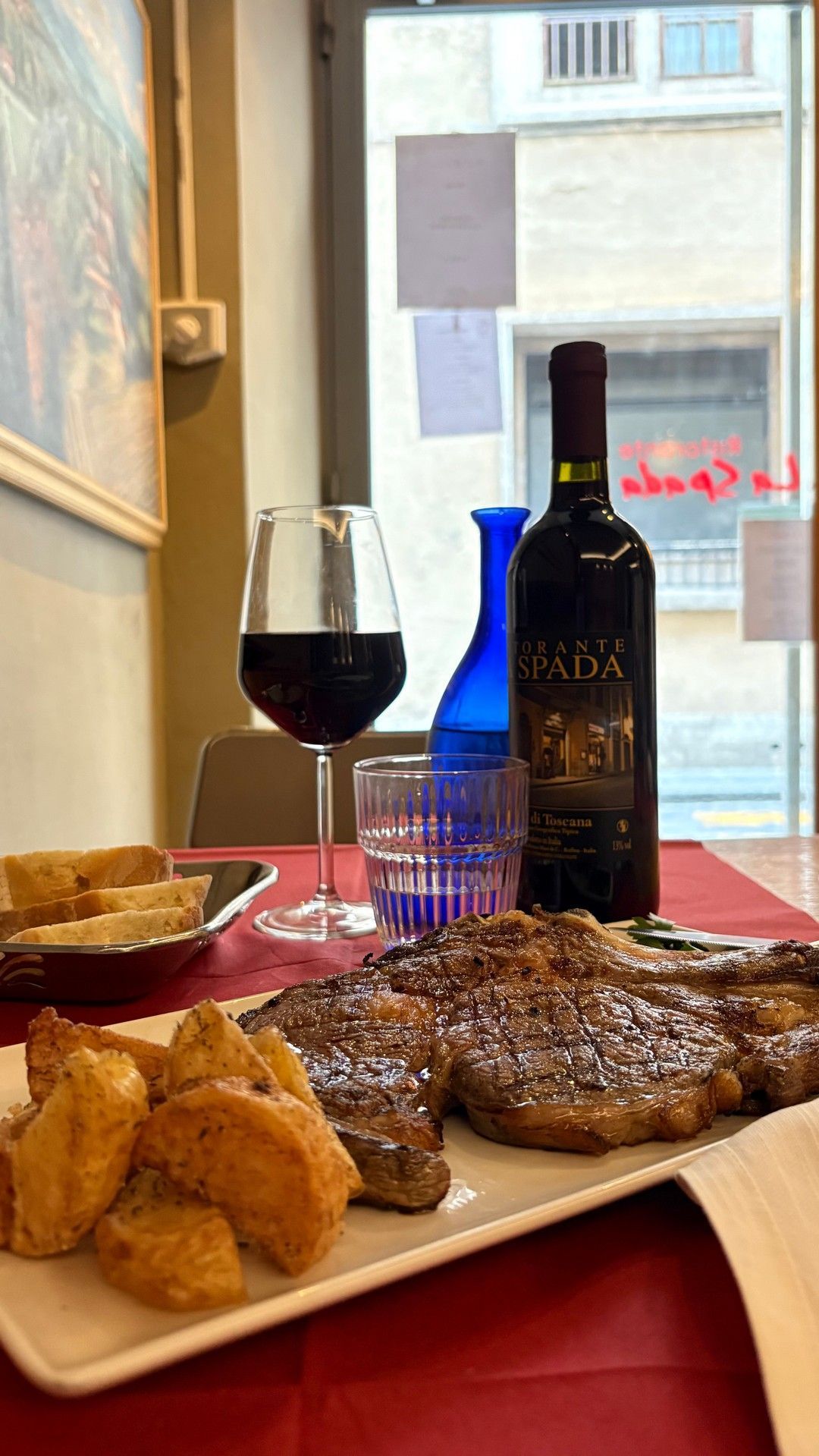 Cena a base di bistecca, vino, patate e vista dalla finestra. Tovaglia rossa, piatto bianco e un vaso blu.