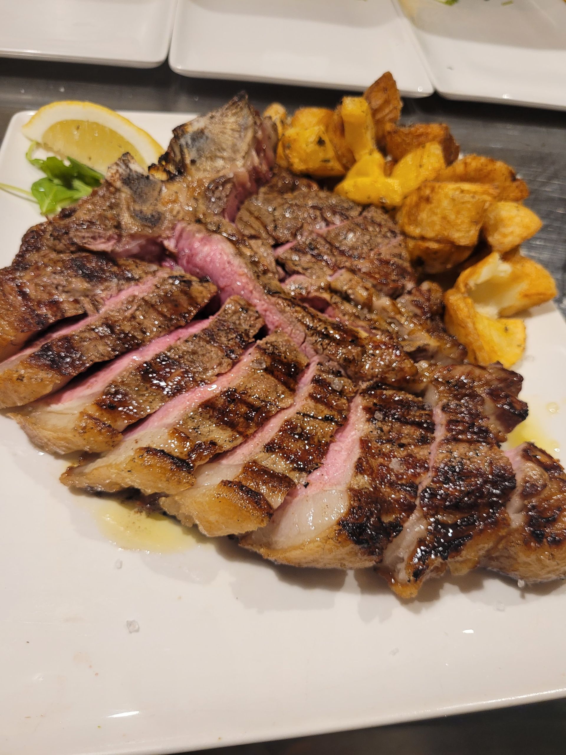 Bistecca grigliata e tagliata a fette con interno rosa, patate, spicchio di limone e rucola su un piatto bianco.