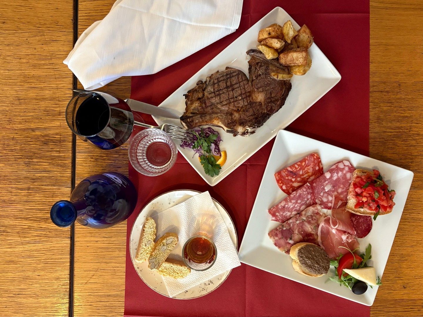 Bistecca, salumi e pane su un tavolo con vino rosso, una brocca blu e un tovagliolo bianco.