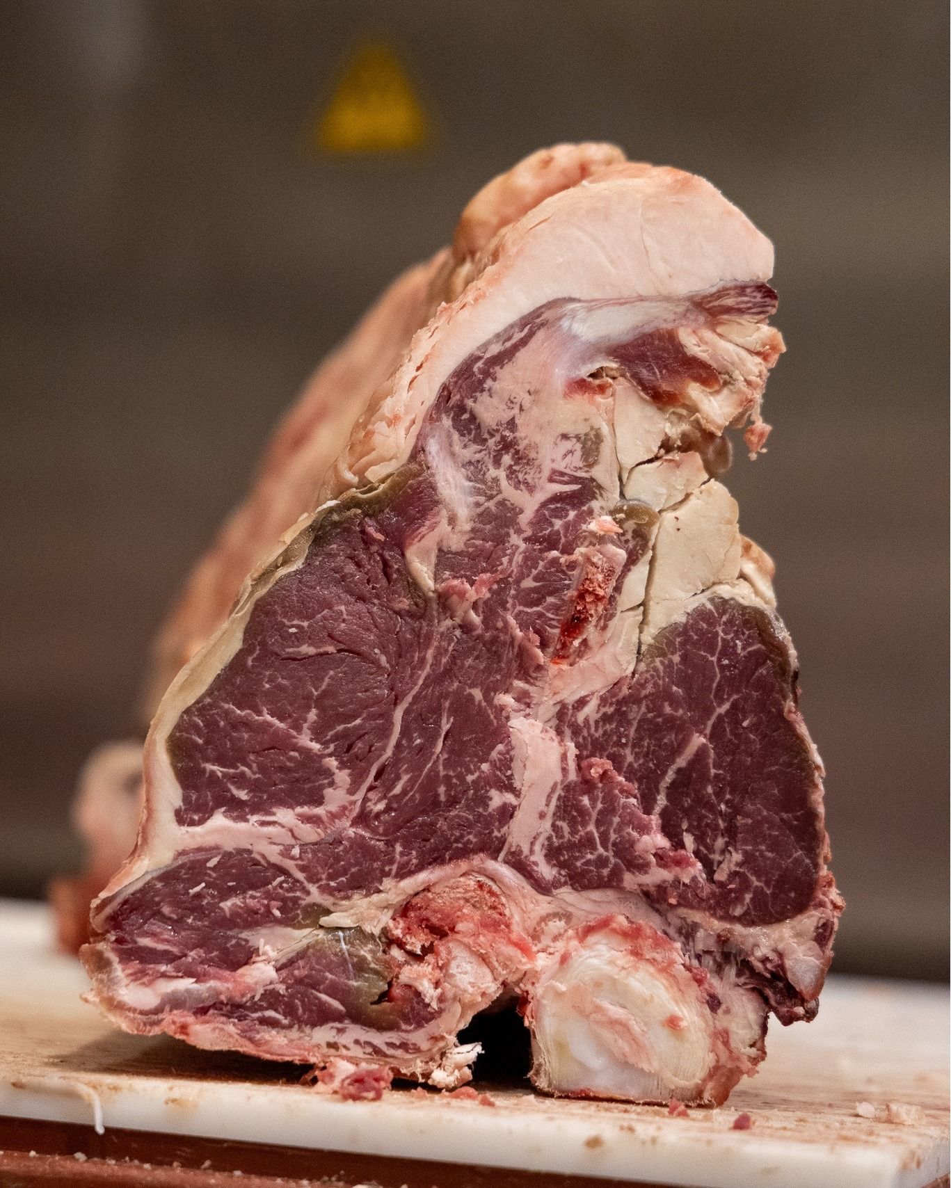 Una grande bistecca T-bone cruda, con osso e marezzatura, appoggiata su un tagliere bianco.