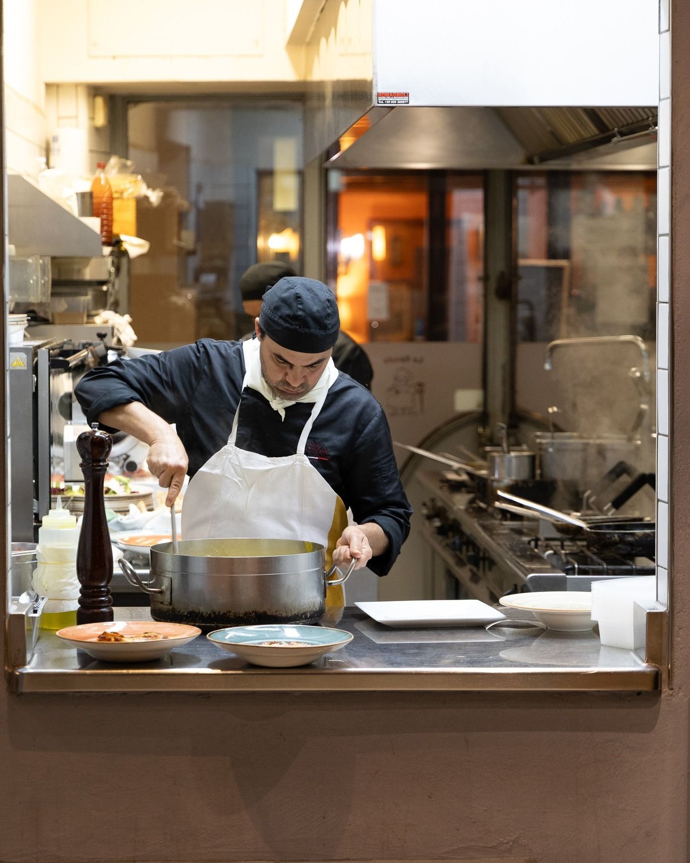 Uno chef che mescola una pentola nella cucina di un ristorante affollato; indossa grembiule e cappello.