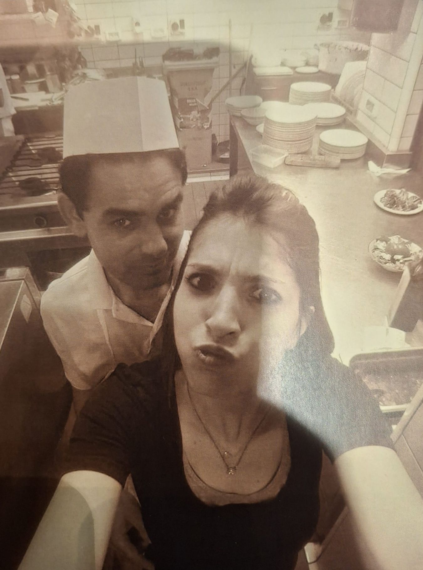 Una donna si scatta un selfie con lo chef in cucina. Lo chef è dietro di lei. Entrambi hanno un'espressione neutra.