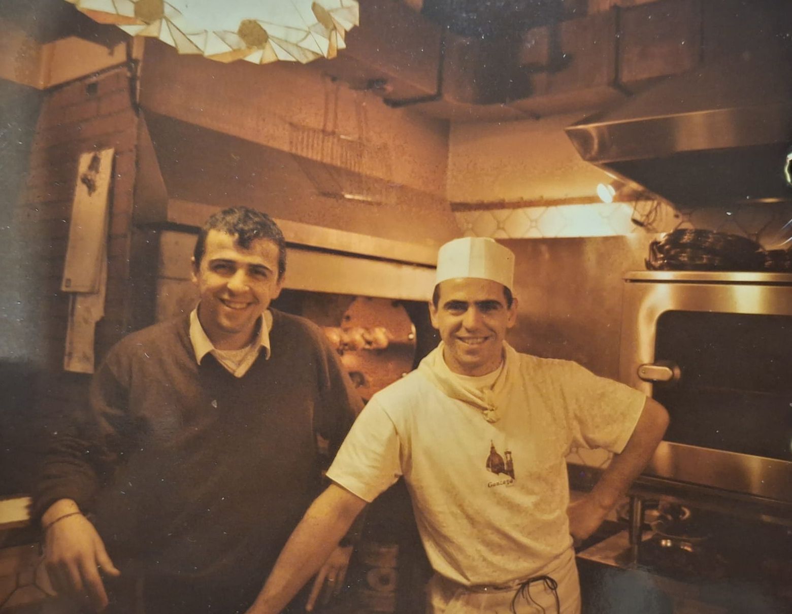 Due uomini sorridenti in una pizzeria, uno in uniforme vicino al forno.