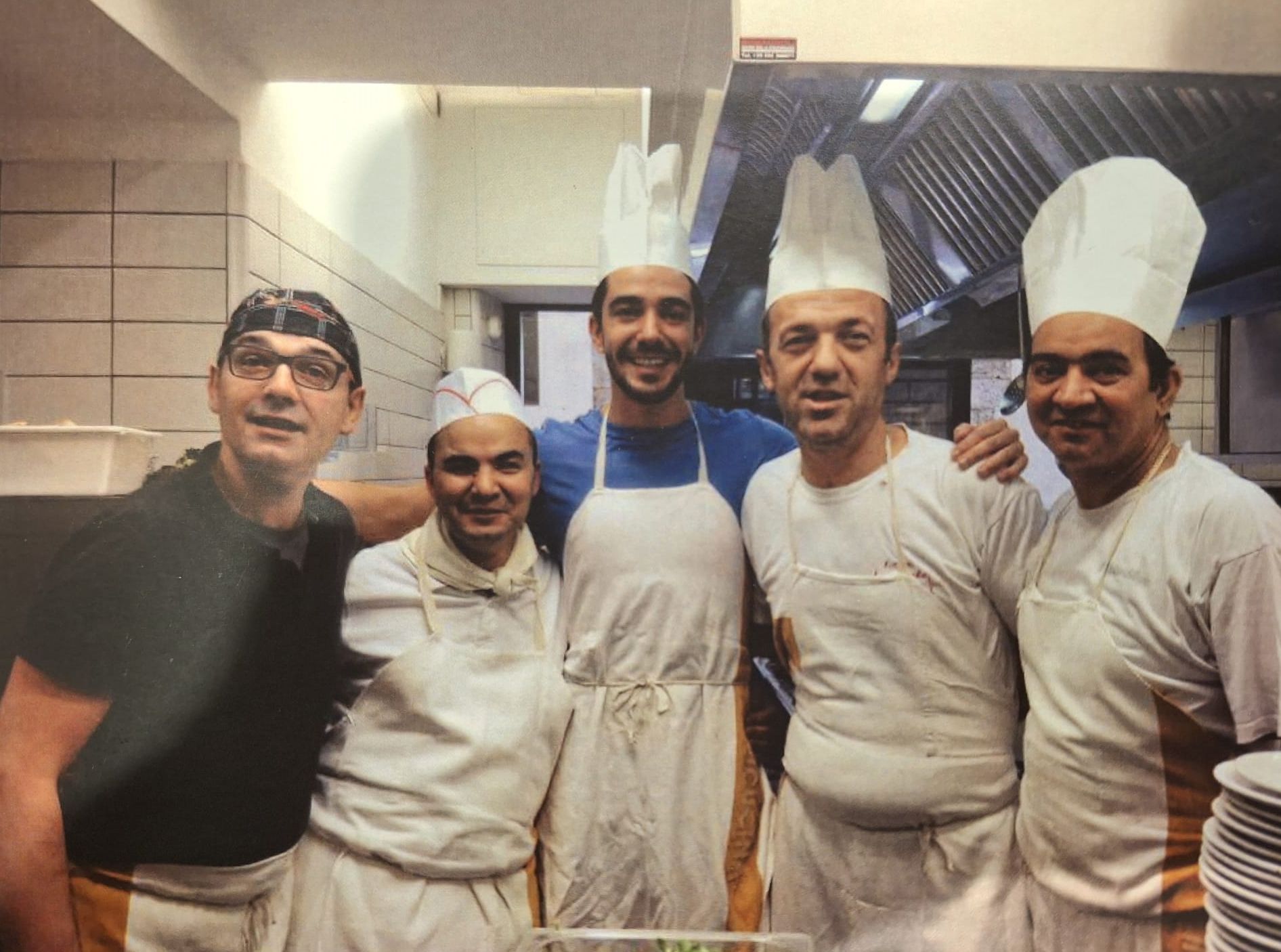 Cinque chef con grembiuli e cappelli bianchi sorridono in cucina.