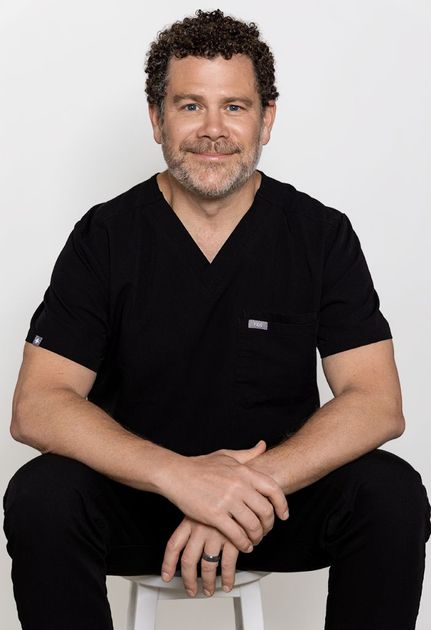 Smiling person in black scrubs seated on a stool against a light gray background.