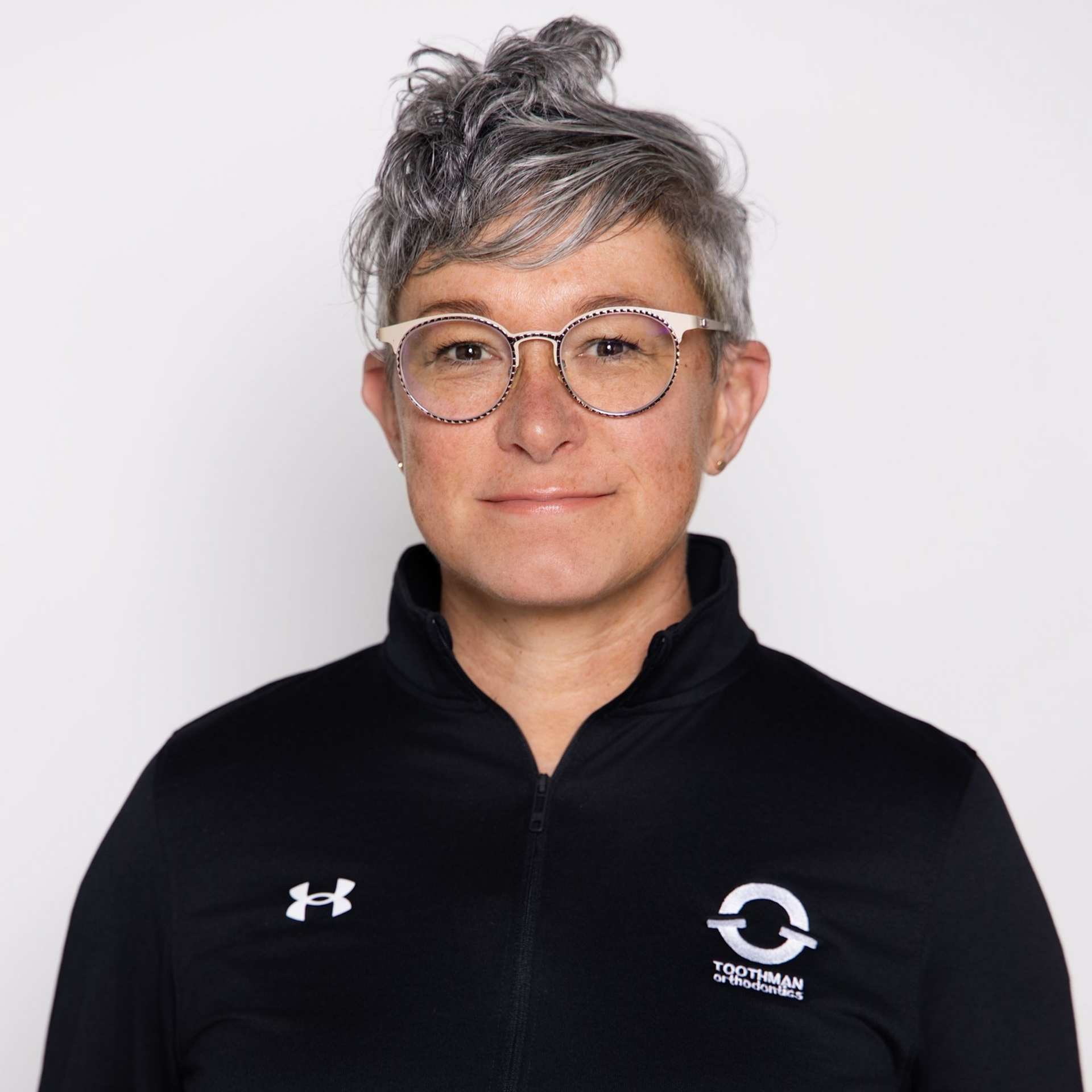 Portrait of a person with short gray hair and glasses in a black zip-up top on a plain background