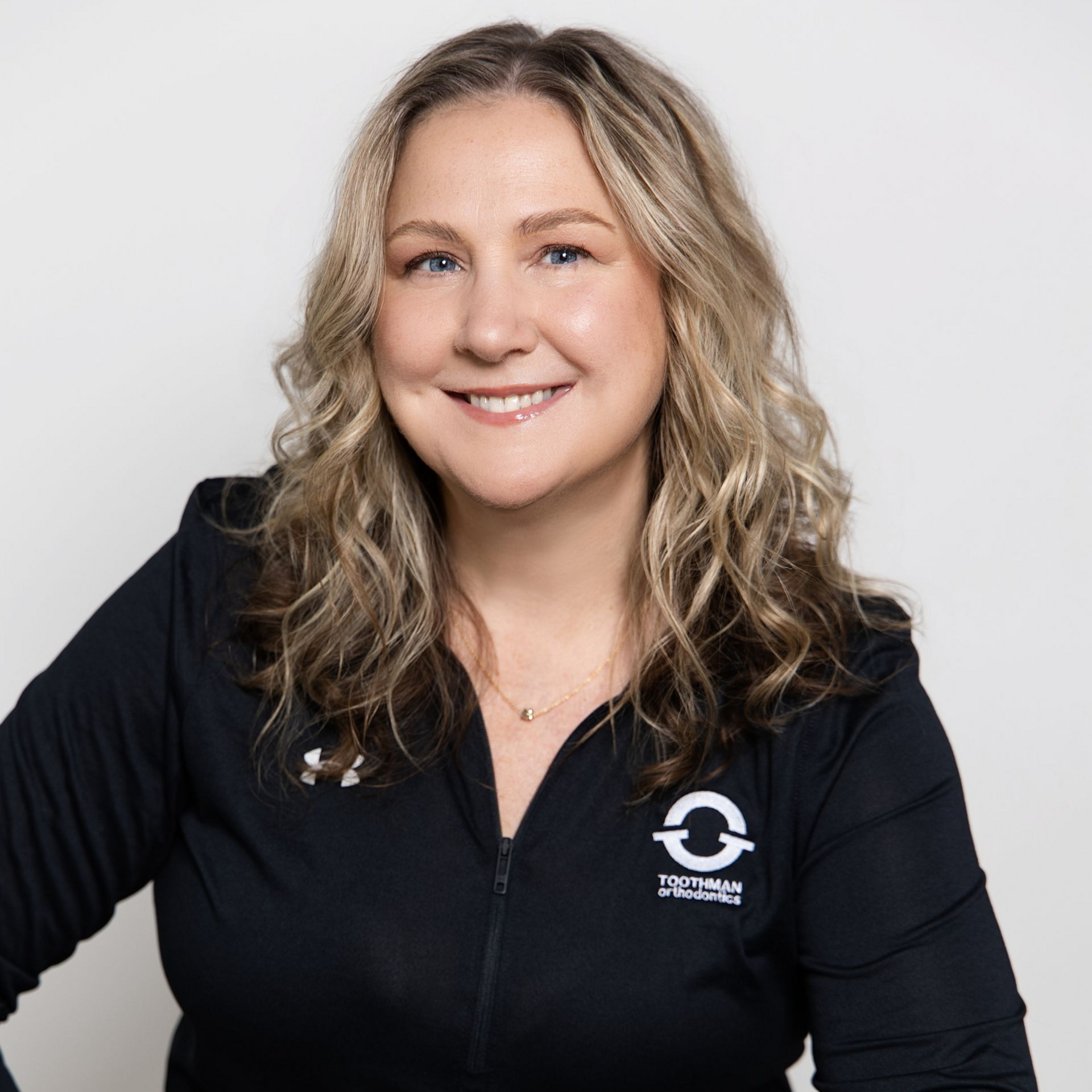 Smiling person with wavy blonde hair in a black zip-up shirt against a light gray background