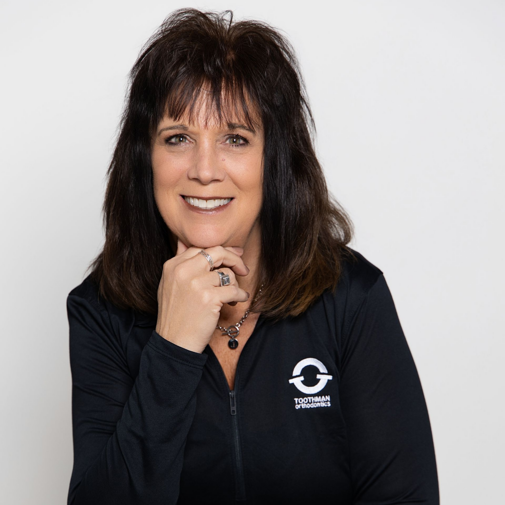 Woman with dark hair in a black top and jeans, seated with hand on chin against a light background