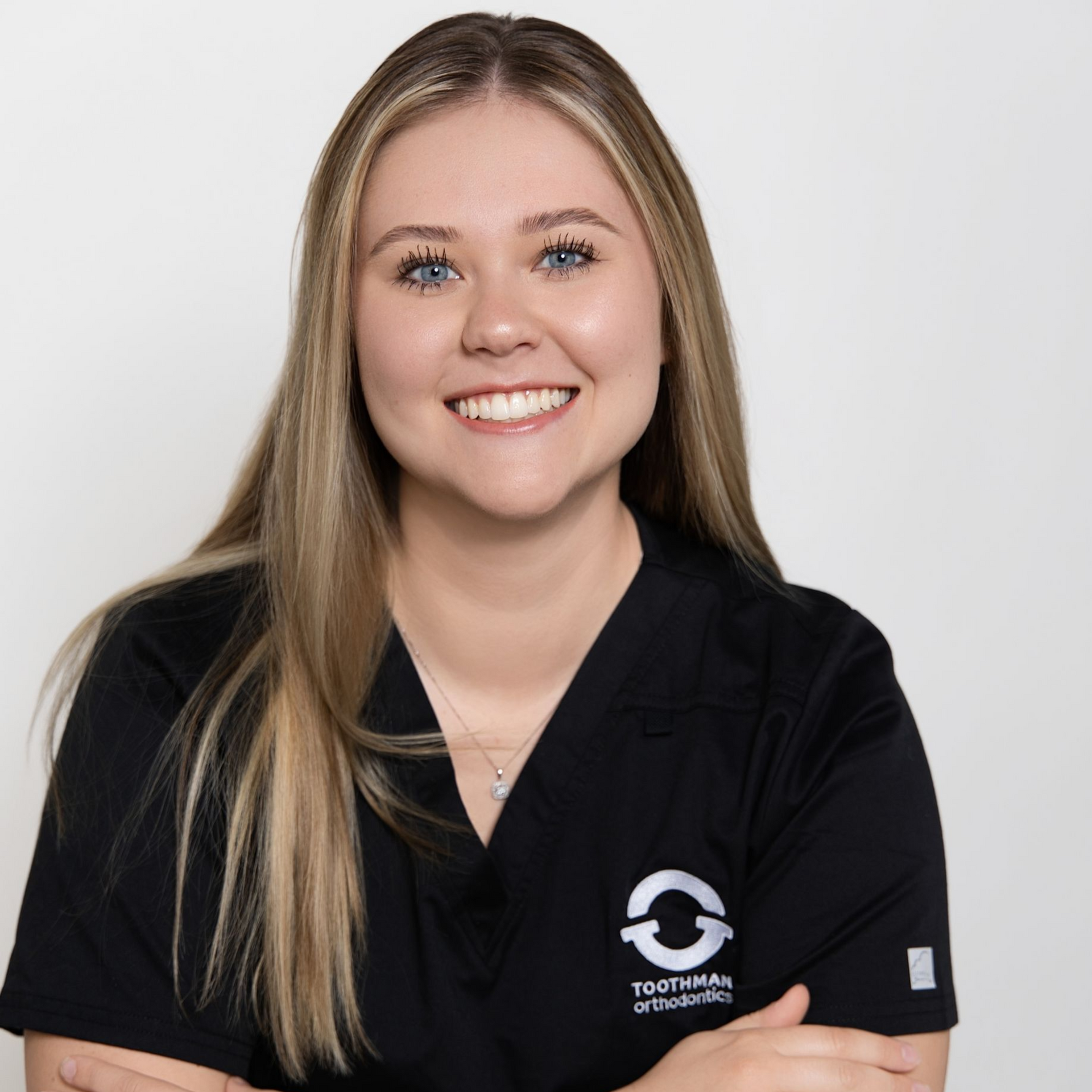Smiling person in black scrubs kneeling with arms crossed against a light gray background