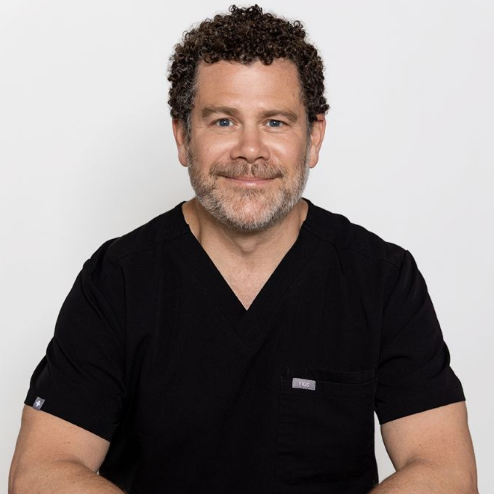 Person seated on a stool, wearing black scrubs and smiling against a plain light background